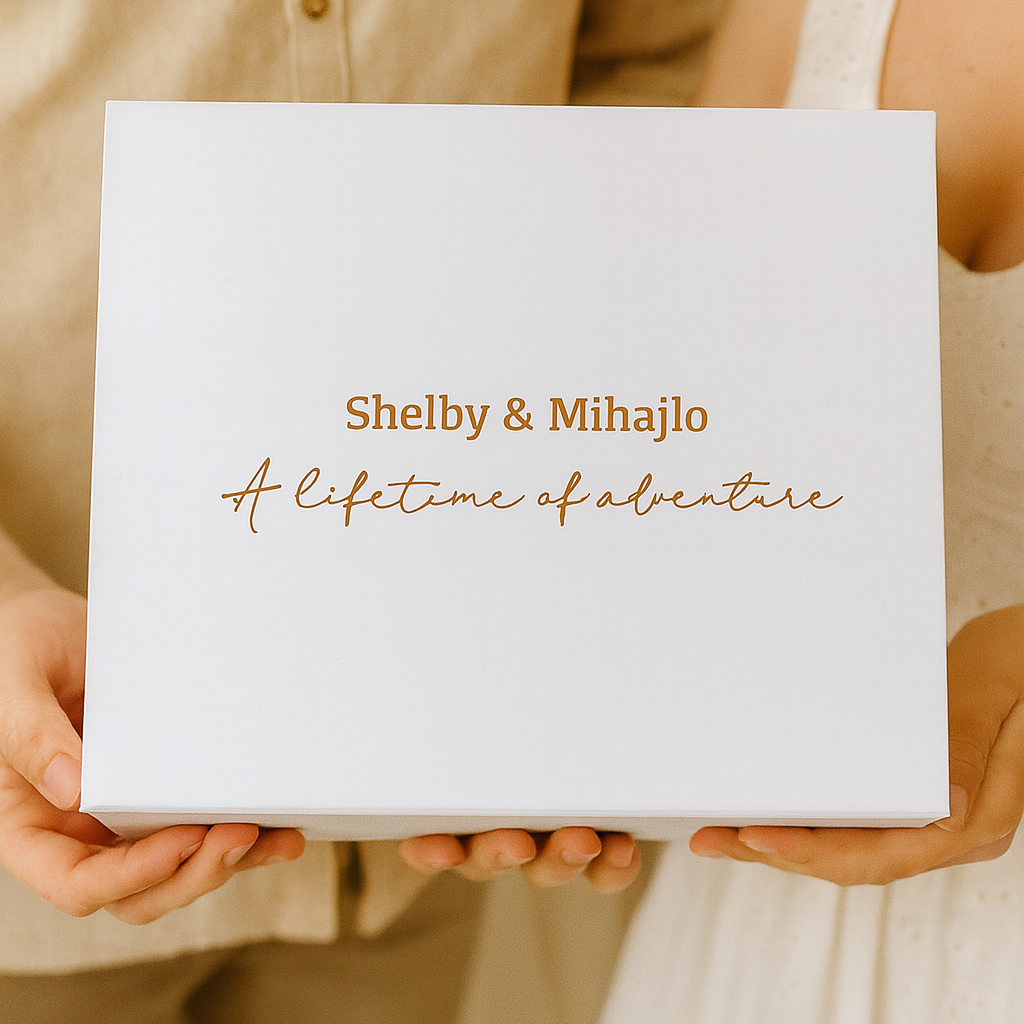Two people holding a white box with gold text 'Shelby & Mihajlo A lifetime of adventure'.