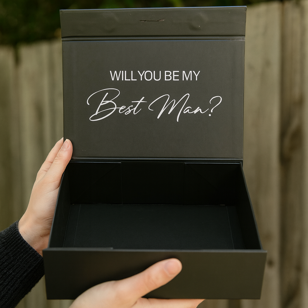 Black box with 'Will You Be My Best Man?' text held by a person outdoors.