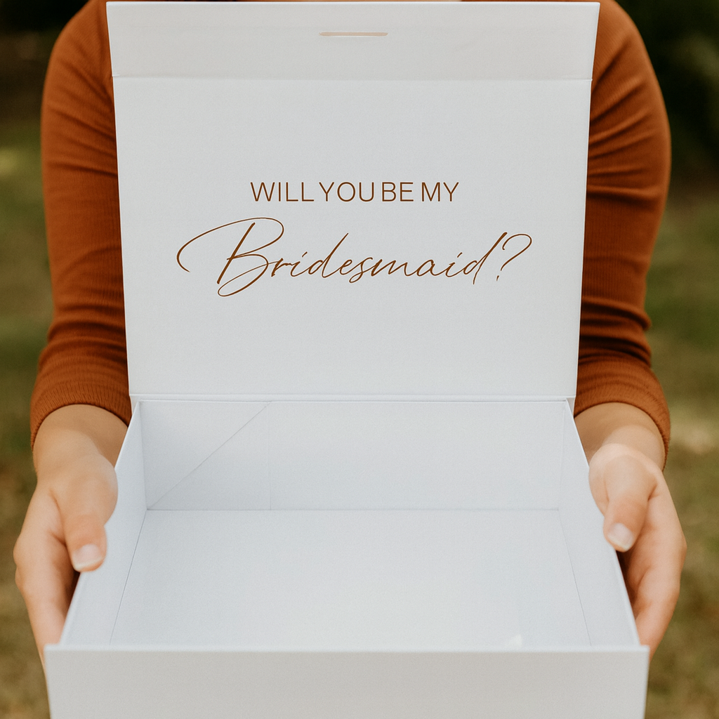Person holding a box with 'WILLYOUBEMY Bridesmaid?' text outdoors.