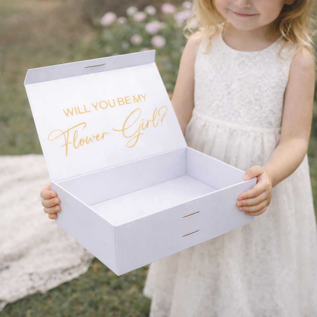 White box with 'Will you be my Flower Girl?' text held by a young girl in a white dress outdoors.