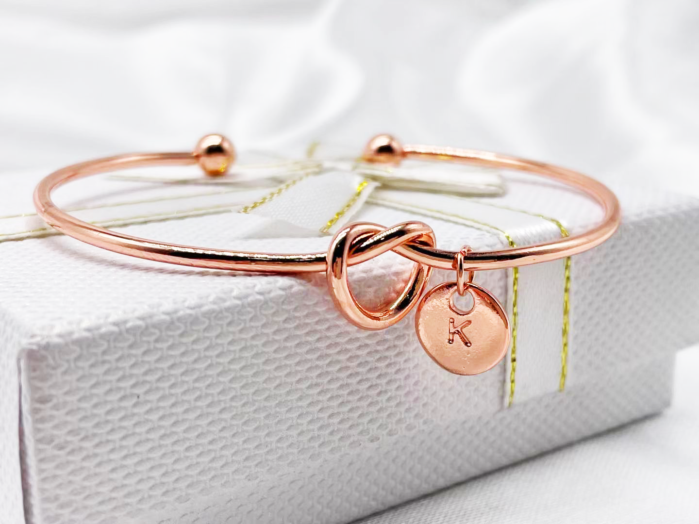 A rose gold-plated bracelet with a personalized initial charm, displayed on a white box with a yellow ribbon.